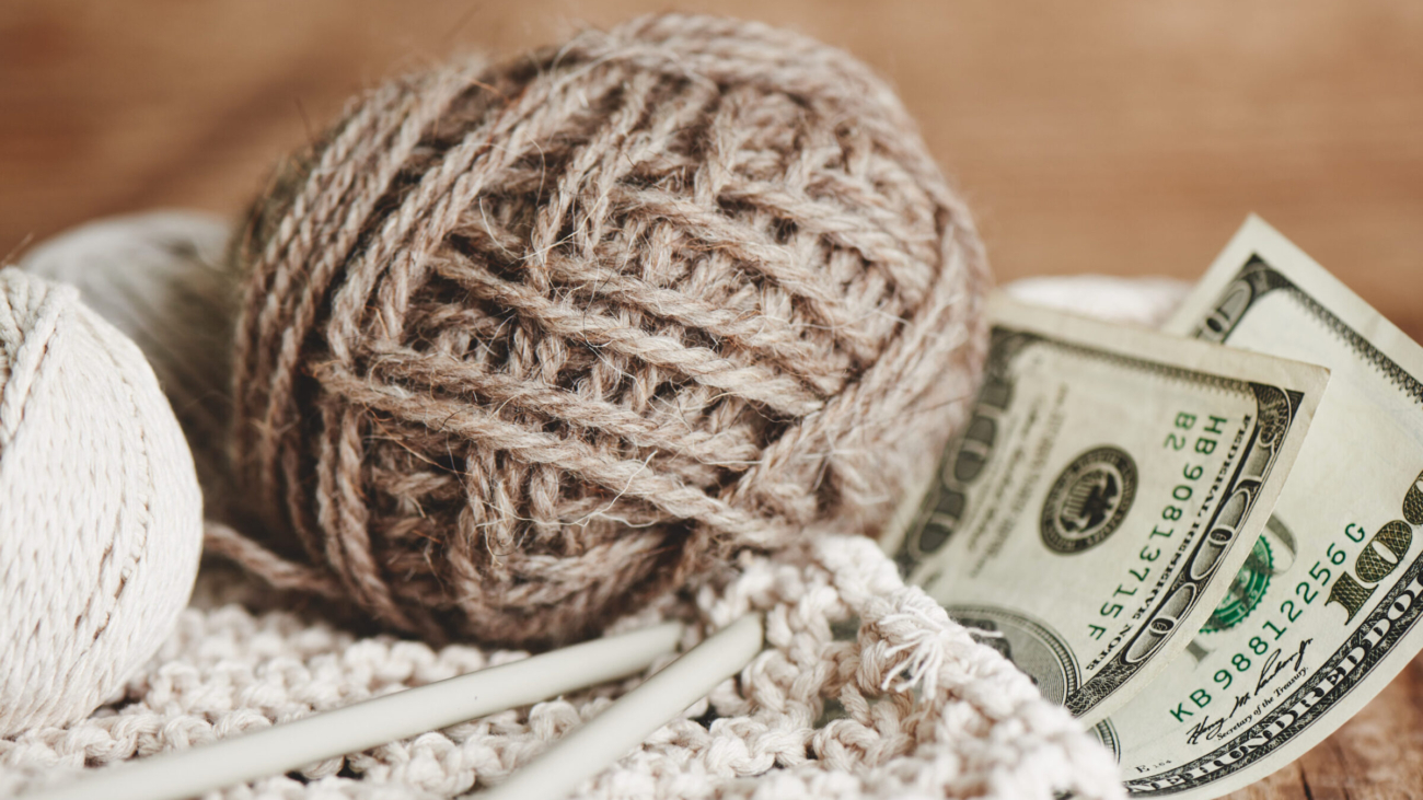 Profitable hobby. Earnings on needlework. Balls of natural color yarn, knitting needles and money on a wooden table.