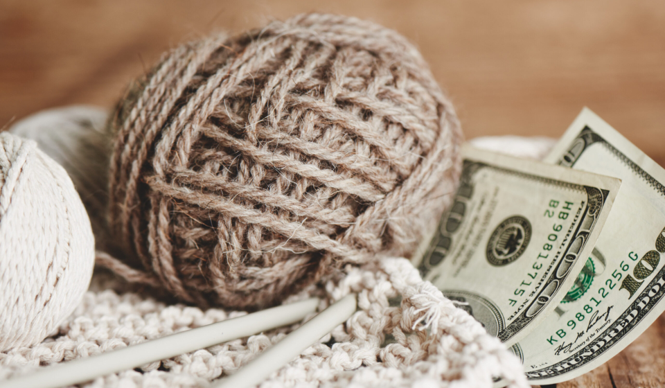 Profitable hobby. Earnings on needlework. Balls of natural color yarn, knitting needles and money on a wooden table.