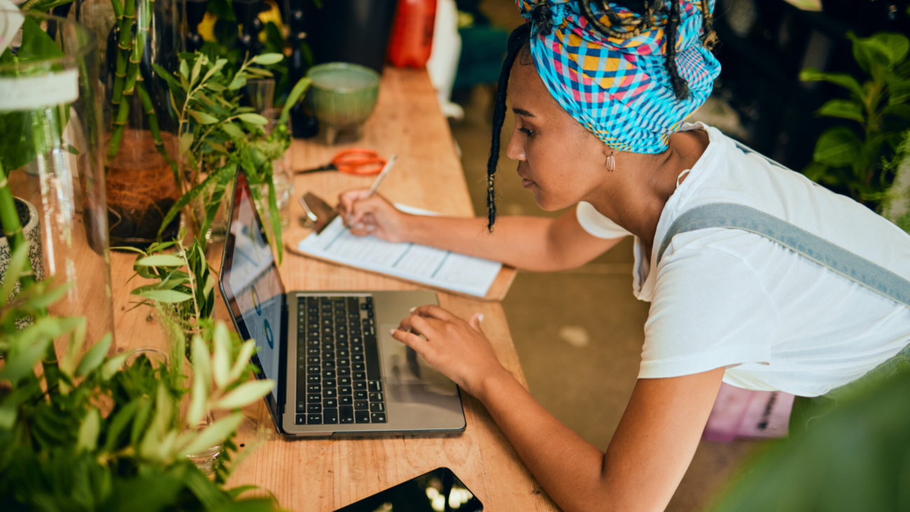 Laptop, small business or black woman writing a checklist on plants or flowers for commerce or stock inventory. Management, store manager or entrepreneur planning or working on floral growth research.