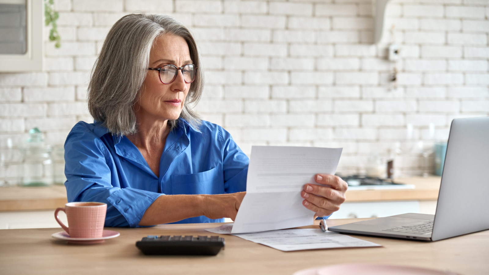 Adult senior 60s woman working at home at laptop. Serious middle aged woman at table holding document calculating bank loan payments, taxes, fees, retirement finances online with computer technologies