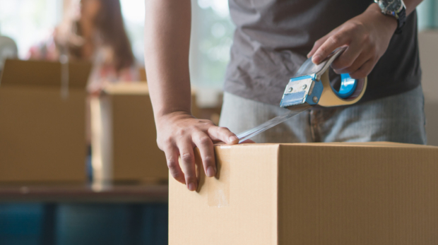Concept young couple moving house. Close-up hand of man use tape sealing cardboard box.