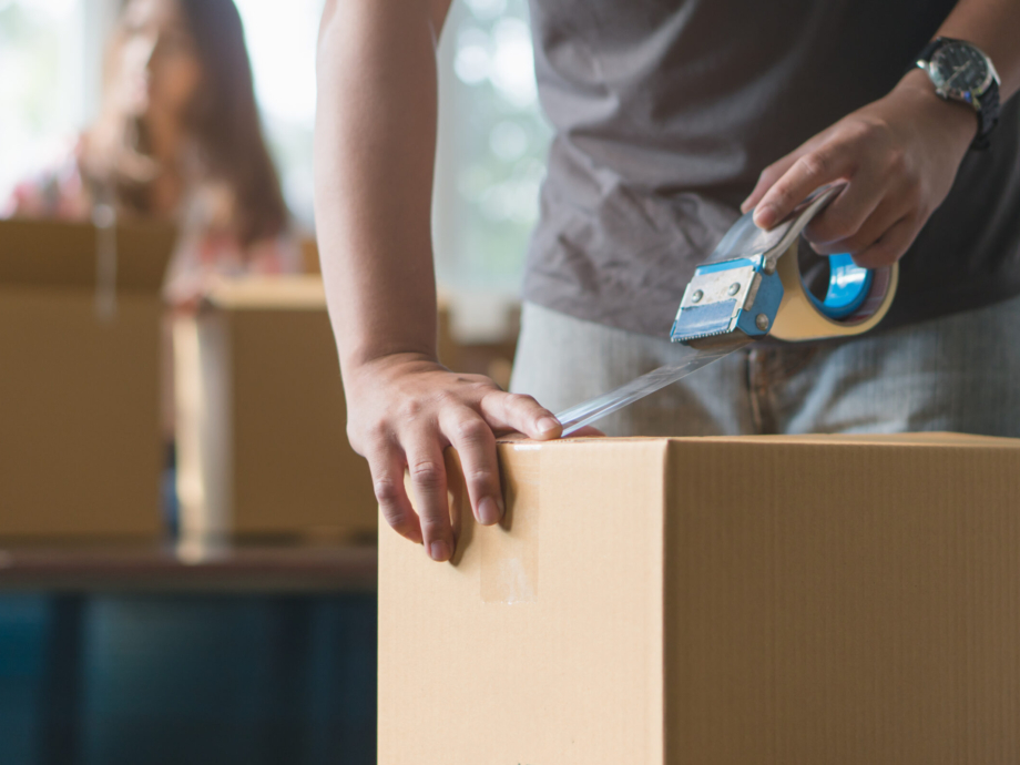 Concept young couple moving house. Close-up hand of man use tape sealing cardboard box.