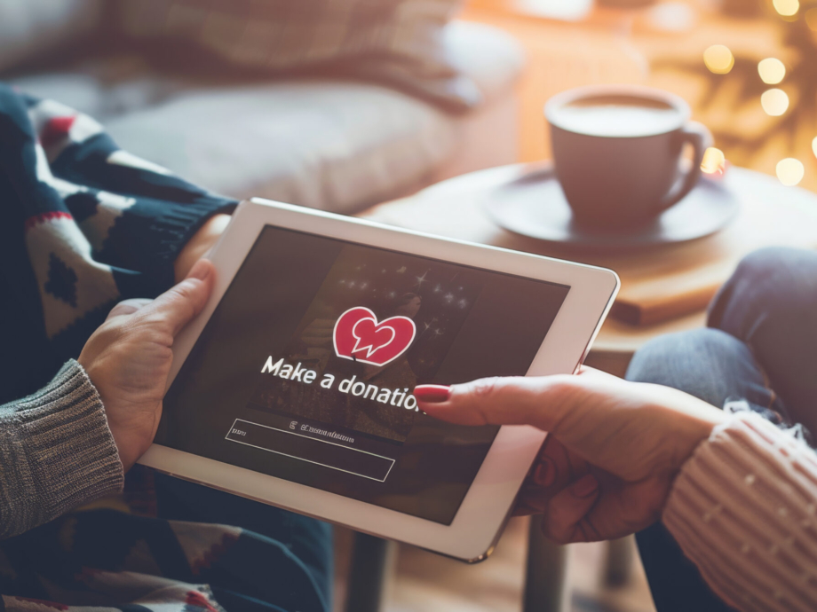 Two hands are holding a tablet displaying a 'Make a donation' screen with a heart symbol, suggesting an online charitable contribution.